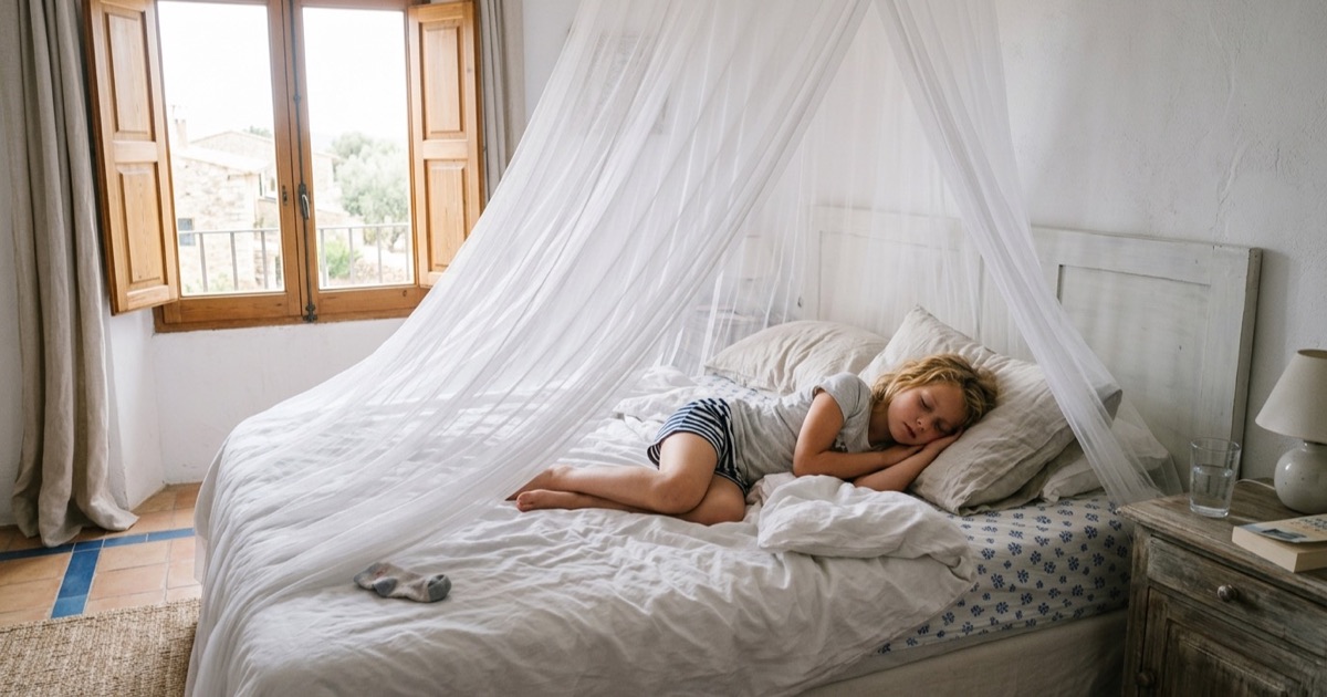 Kind ligt onder een muskietennet op een bed in een vakantiehuis met daglicht door open ramen.
