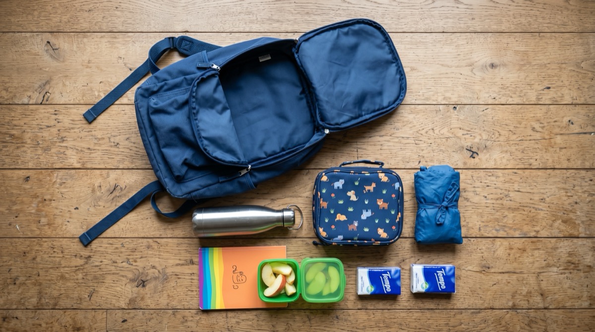 Een geopende blauwe kinderrugzak met inhoud op houten vloer: broodtrommel, drinkbeker, notitieboek en zakdoeken.
