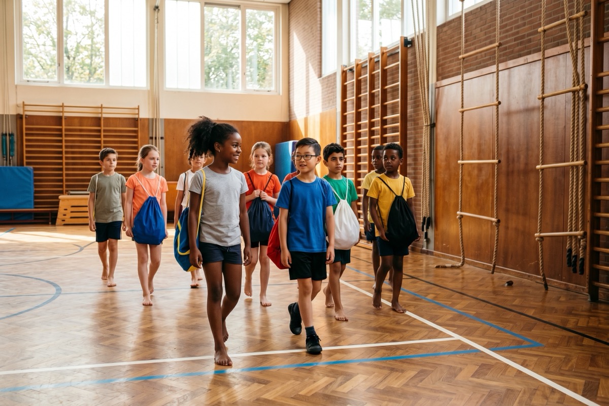 Diverse groep basisschool-kinderen in eenvoudige gymkleding loopt met gymtassen door een gymzaal met houten vloer en wandrek.